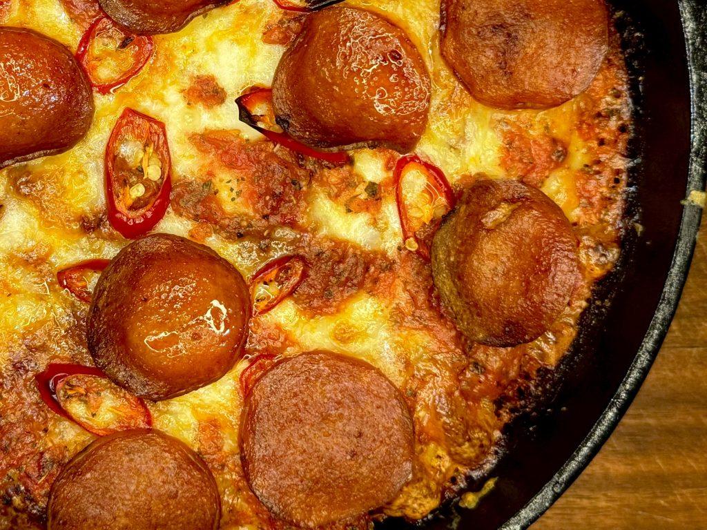 Cast iron pan on wooden board with a crispy, fluffy pepperoni pan pizza in it.