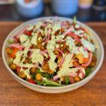 Bowl on wooden board containing salad, charred red peppers, massala chickpeas and pickled red onion with green chutney dressing.