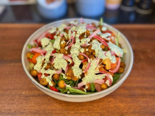 Bowl on wooden board containing salad, charred red peppers, massala chickpeas and pickled red onion with green chutney dressing.