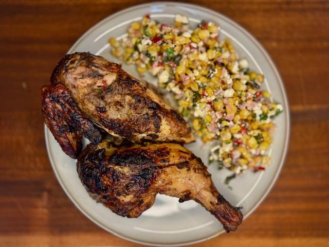 Plate on wooden board with half a BBQ jerk chicken and grilled corn, feta and chilli salad.