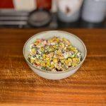 Bowl on wooden board containing salad of grilled corn, red chilli, feta cheese and herbs with lime.