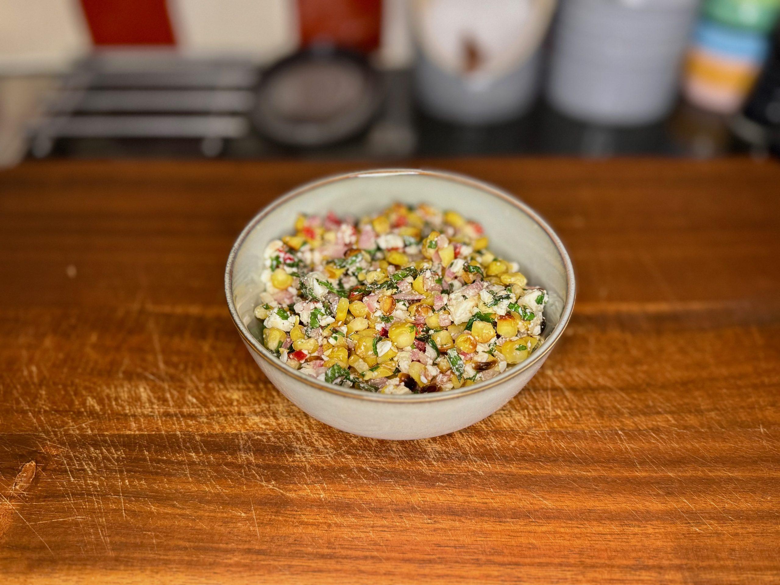 Bowl on wooden board containing salad of grilled corn, red chilli, feta cheese and herbs with lime.