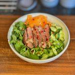 Bowl of salad on a wooden board containing vegetables, herbs, noodles and steak with a vietnamese dressing.