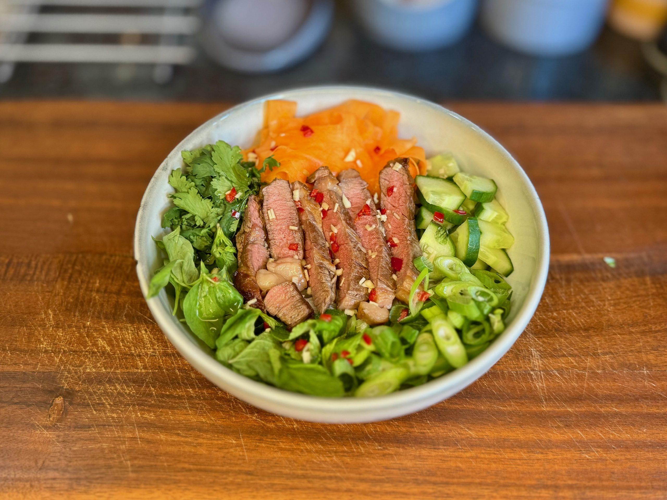 Bowl of salad on a wooden board containing vegetables, herbs, noodles and steak with a vietnamese dressing.