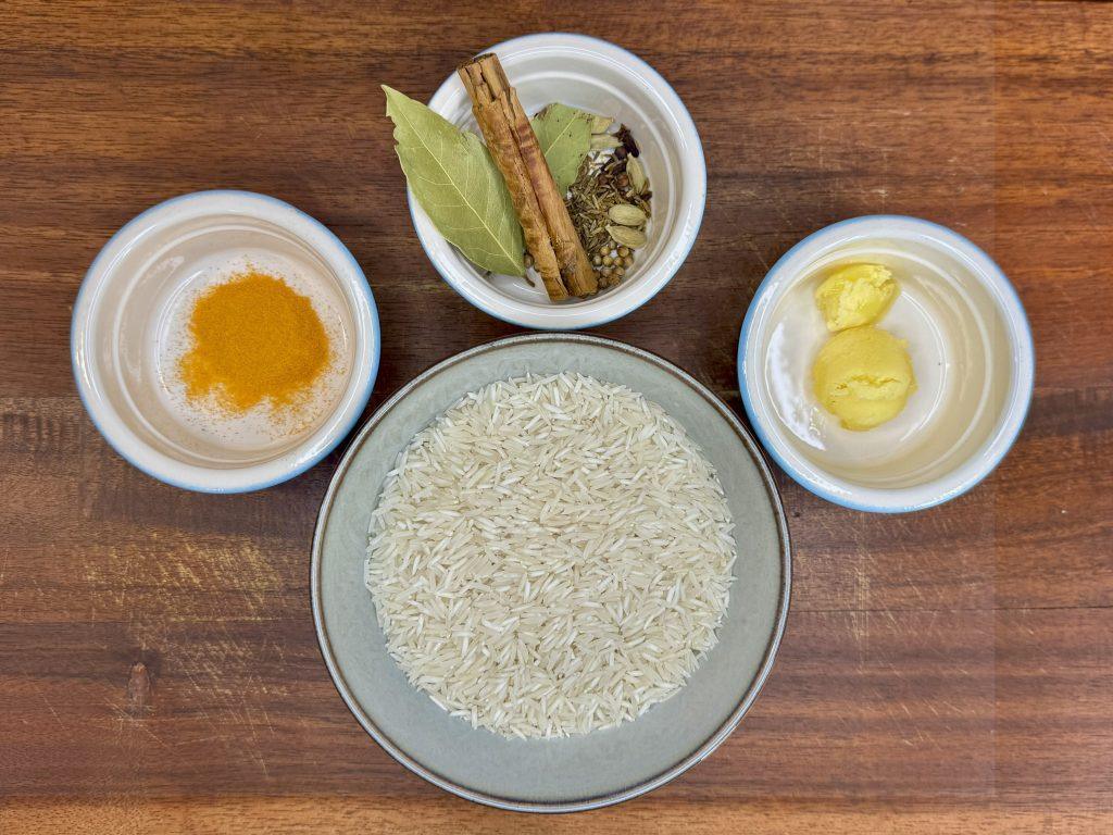 A bowl of rice, bow of ghee, bowl of turmeric and bowl of whole spices on a wooden board.