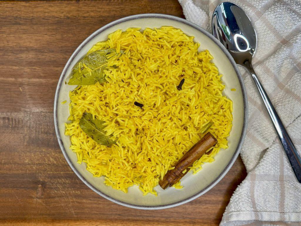 Bowl of yellow pilau rice with bay leaf and cinnamon stick. Bowl sitting on wooden chopping board next to a towel and a spoon.
