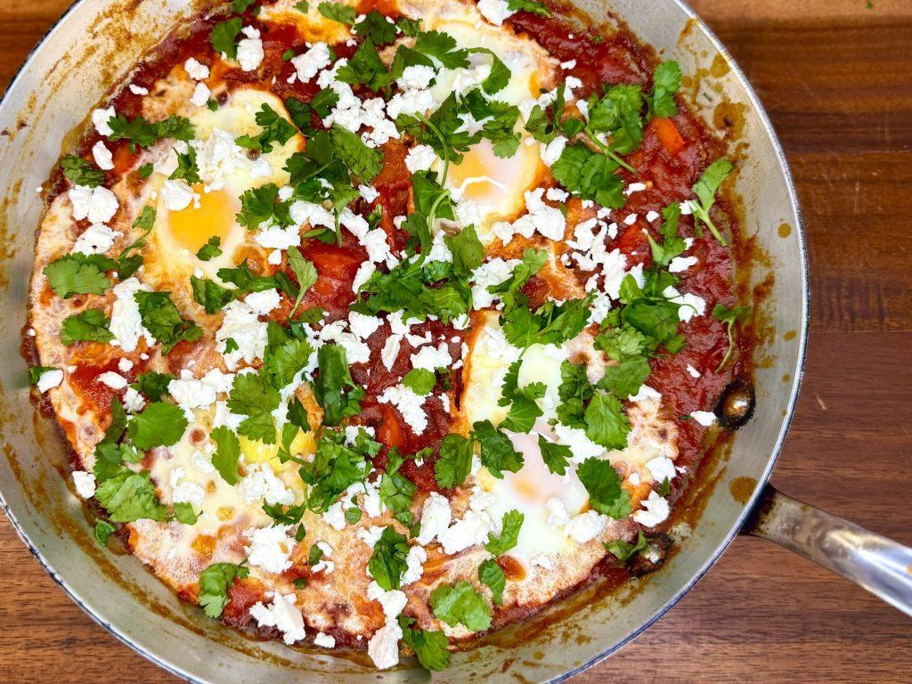Pan of shakshuka on a wooden chopping board with feta and coriander garnishing it.