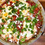 Pan of shakshuka on a wooden chopping board with feta and coriander garnishing it.