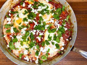 Pan of shakshuka on a wooden chopping board with feta and coriander garnishing it.