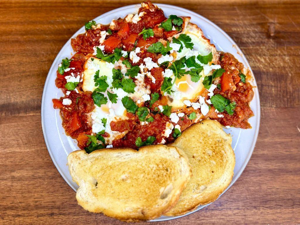 Plate of shakshuka on a wooden chopping board. Garnished with feta and coriander and served with toast.