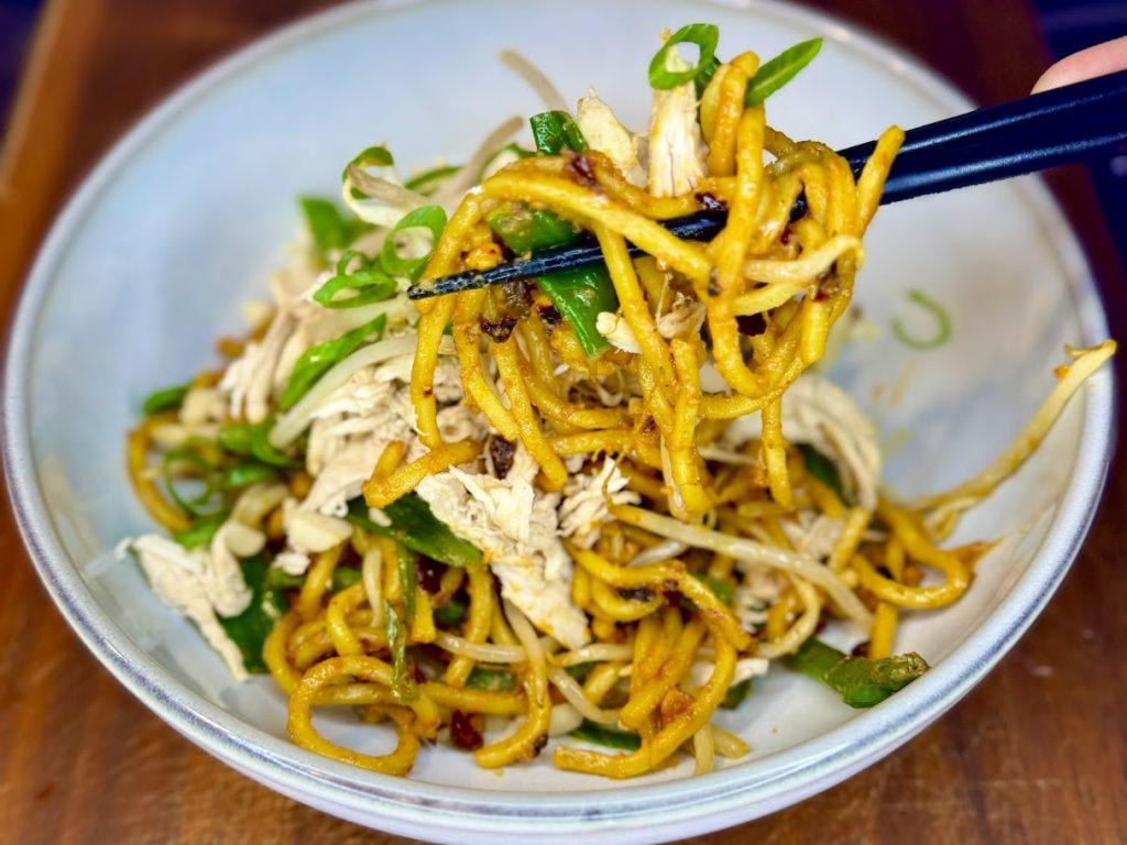 Bowl of peanut and chilli oil noodles with shredded, poached chicken on a wooden board being lifted with black chopsticks.