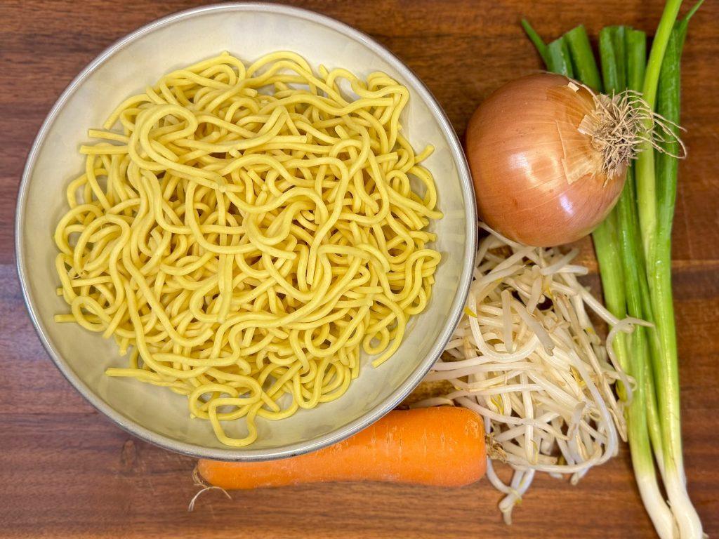 Noodles, an onion, a carrot and spring onions on a wooden board.