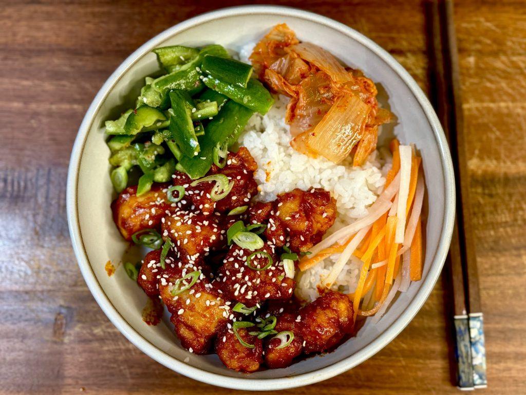 Bowl of crispy korean fried chicken, rice, kimchi, pickles and cucumber with chopsticks