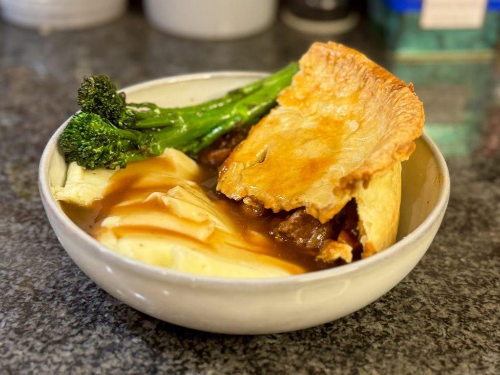 A slice of steak and ale pie in a bowl with mashed potatoes, broccoli and gravy