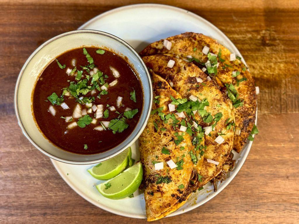 Birris tacos with a bowl of birria sauce and sliced limes.