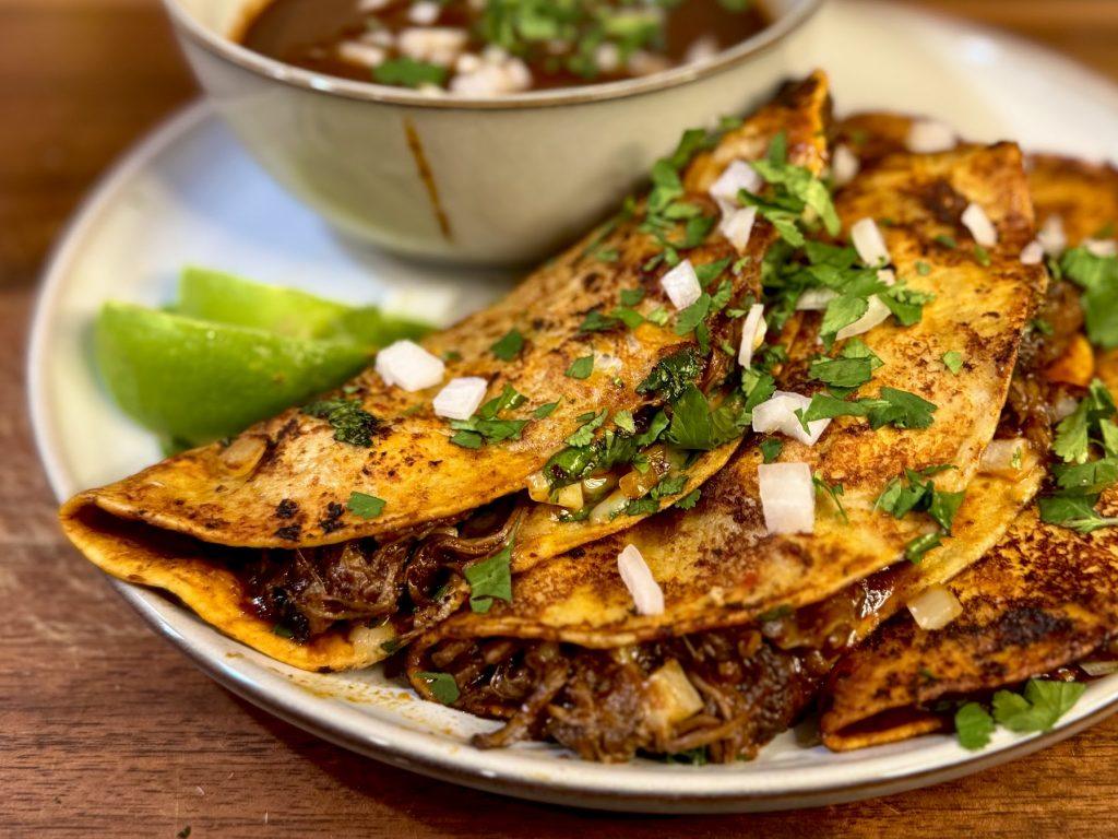 Birris tacos with a bowl of birria sauce and sliced limes.