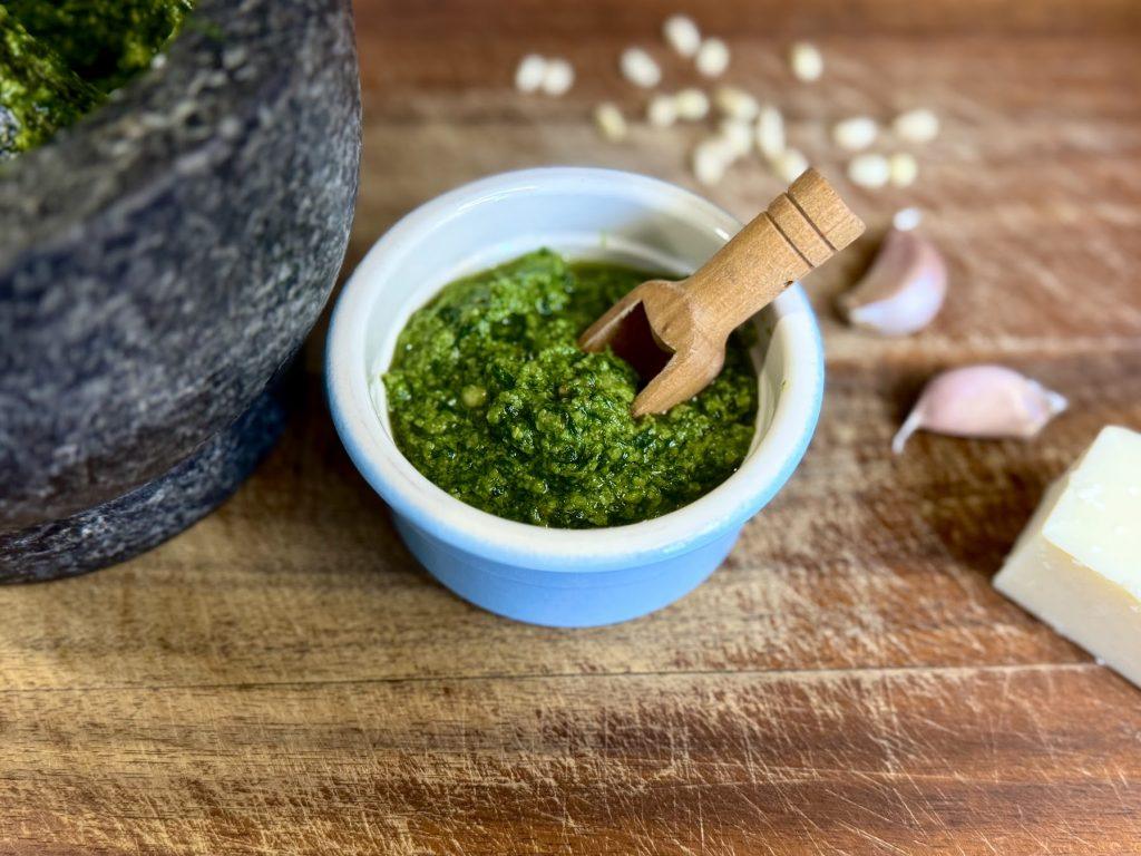 A bowl of pesto alla genovese and a wooden spoon on a board with ingredients